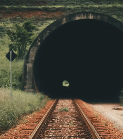 israel-oliveira-XAt2el_iwyg-unsplash Railway tracks leading into a dark tunnel surrounded by greenery.